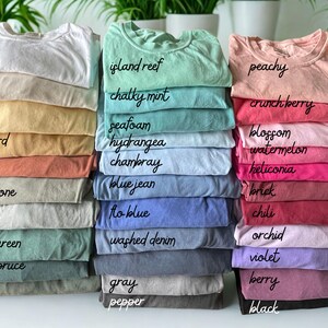 May include: Stacks of folded t-shirts in various colors, including white, peachy, and blue jean. Each stack is labeled with the color name in cursive script. The shirts are neatly arranged, showcasing a range of hues.