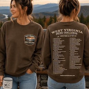 May include: Two women wearing brown sweatshirts. The front of the sweatshirt has a patch with the text "WEST VIRGINIA SWING CHALLENGE" and a mountain scene. The back of the sweatshirt has a list of locations. One woman holds a mug that says "LIFE IS BETTER IN THE MOUNTAINS".