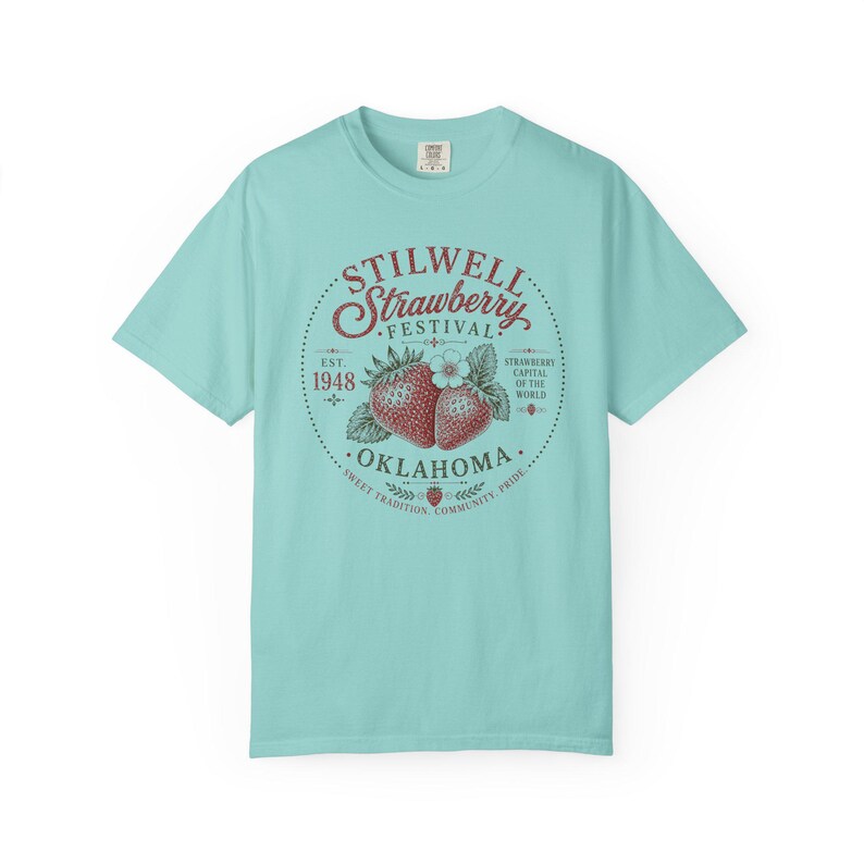 May include: A light blue t-shirt featuring a circular graphic for the Stilwell Strawberry Festival. The graphic includes the text "STILWELL Strawberry FESTIVAL" and "OKLAHOMA" with illustrations of strawberries and a flower. The text "EST. 1948" is also present.
