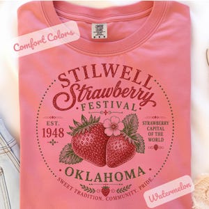 May include: A pink t-shirt with a graphic design for the Stilwell Strawberry Festival in Oklahoma. The design includes strawberries, text, and the words "Strawberry Capital of the World". The shirt also has the words "Comfort Colors" and "Watermelon".