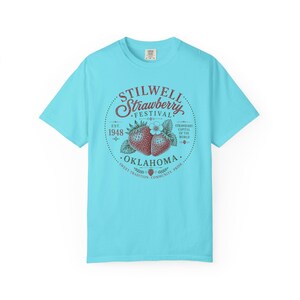 May include: A light blue t-shirt featuring a circular graphic with the text "STILWELL STRAWBERRY FESTIVAL" and "OKLAHOMA". The graphic includes illustrations of strawberries and the text "EST. 1948".