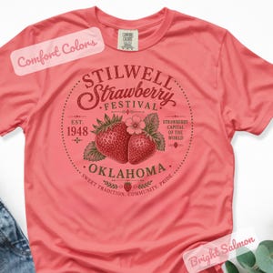 May include: A bright salmon-colored t-shirt featuring a circular design with the text "STILWELL Strawberry Festival OKLAHOMA" and illustrations of strawberries. The shirt also includes the text "EST. 1948" and "Strawberry Capital of the World."