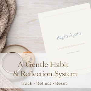 Puede incluir: Una manta beige suave y una taza de café con una galleta se encuentran junto a un diario titulado "Begin Again". El diario es parte de "A Gentle Habit & Reflection System" con las palabras "Track, Reflect, Reset" debajo.