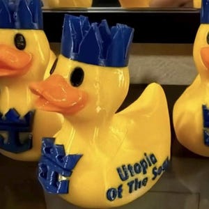 May include: A yellow rubber duck featuring an orange beak, a blue crown, and a blue anchor emblem on its chest. The side of the duck displays the blue text Utopia Of The Seas. The duck is part of a group of identical items shown in the image.
