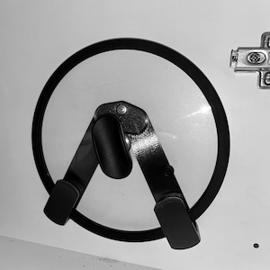 May include: Black metal pot lid holder mounted on a white surface. The holder has a circular frame and two angled arms with rubberized grips to secure the lid. A clear glass lid is held in place. A metal hinge is visible.