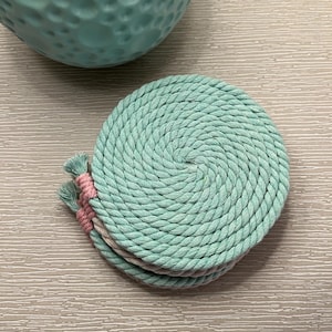 Puede incluir: Un conjunto de cuatro posavasos redondos hechos de cuerda en espiral de color verde azulado claro, con detalles de borlas rosas. Un cuenco de cerámica texturizada, de color verde azulado, está en el fondo. Los posavasos están sobre una superficie gris clara.