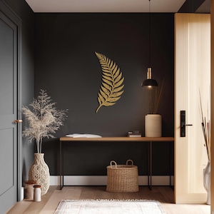 Könnte beinhalten: Goldfarbene Farnblatt-Wanddekoration an einer dunkelgrauen Wand. Der Raum verfügt über einen Holztisch, einen Weidenkorb und eine beige Vase mit getrockneten Pflanzen. Eine schwarze Pendelleuchte hängt über dem Tisch.