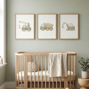 Könnte beinhalten: Drei gerahmte Drucke von Baumaschinen: ein Kran, ein Muldenkipper und ein Bagger. Die Drucke befinden sich in einem Kinderzimmer, über einem Holzbettchen mit einer cremefarbenen Decke und einem runden Kissen. Eine kleine Pflanze und ein geflochtener Korb mit Holzklötzen sind ebenfalls zu sehen.