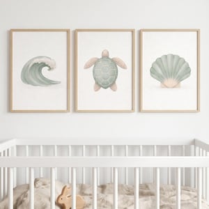 Könnte beinhalten: Drei gerahmte Aquarelldrucke mit Meeresthemen. Die Drucke zeigen eine Welle, eine Meeresschildkröte und eine Muschel, alle in sanften Grün- und Beigetönen. Das Kunstwerk wird über einem weißen Kinderbett mit einem Holzkaninchenspielzeug ausgestellt.