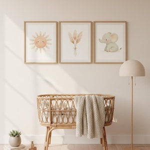 May include: Three framed watercolor prints with a nursery theme. The prints feature a smiling sun, pampas grass, and a baby elephant. A woven bassinet with a blanket, a floor lamp, and a small potted plant complete the nursery decor.