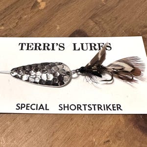May include: A fishing lure with a hammered silver-colored metal body and a feathered tail. The lure is attached to a hook and is on a white card with the text "TERRI'S LURES" and "SPECIAL SHORTSTRIKER".