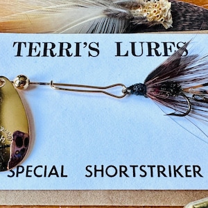 May include: A gold-colored fishing lure with a teardrop-shaped spinner blade and a feathered tail. The lure is attached to a white card with the text "TERRI'S LURES SPECIAL SHORTSTRIKER". Feathers and a wooden surface are in the background.