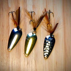 Puede incluir: Tres señuelos de pesca con cuerpos metálicos en forma de lágrima y detalles de plumas. Uno es plateado, otro dorado y el tercero tiene un acabado texturizado y martillado. Cada uno tiene un anzuelo triple.