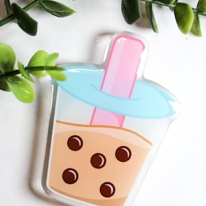 May include: A clear, bubble tea-shaped sticker with a pink straw, light blue lid, and brown tapioca pearls. The drink is a light brown color. Green leaves are in the background.