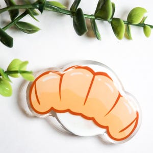 May include: A clear acrylic pin featuring a cartoon croissant design. The croissant is light orange with darker orange lines and a white base. Green leaves are in the background.