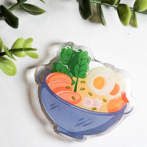 Peut inclure: Un autocollant acrylique transparent représentant un bol de ramen bleu. Les ramen comprennent des crevettes, des œufs, des légumes verts et des nouilles. L'autocollant a une bordure blanche et une plante verte feuillue en arrière-plan.