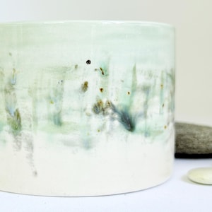 May include: A cylindrical ceramic planter with a white base and a light green top. The planter has a textured, glazed finish with streaks of green, blue, and brown. The planter is approximately 13 cm tall and 13 cm in diameter.