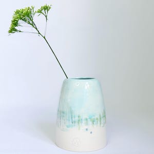 May include: A handcrafted ceramic vase with a light blue and white glaze. The vase has an organic shape and a textured surface. A green stem with small white flowers is placed inside the vase. The vase is approximately 18 cm tall.