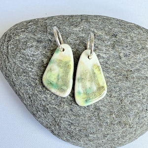May include: A pair of ceramic earrings with silver hoops. The earrings have an irregular shape and are painted with pale yellow, green, and white colours. Small black speckles add detail to the surface. The earrings are handmade.