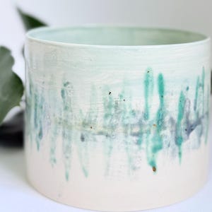May include: A cylindrical ceramic planter with a white base and a pale blue top. The planter has vertical streaks of teal and dark blue paint, creating a textured, artistic design. Suitable for indoor plants.