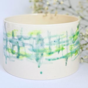 May include: A cream-coloured ceramic bowl with a cylindrical shape. The bowl features a hand-painted design of overlapping blue and green brushstrokes. The interior of the bowl is also cream-coloured.