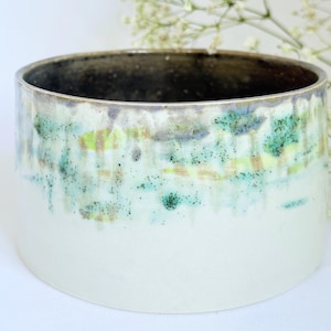 May include: A cylindrical ceramic planter with a white base and a dark interior rim. The upper section has a mix of green, blue, and yellow tones with speckled details. Suitable for indoor plants, this planter is a decorative item.