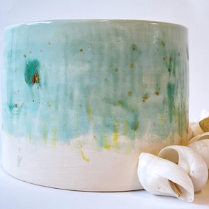 May include: A cylindrical ceramic planter with a white base and a turquoise and green upper section. The planter has a speckled design and is next to a pile of white seashells.