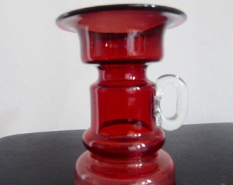 May include: A red glass vase with a wide, flat top and a single handle. The vase has a unique, cylindrical shape with a bulbous base.