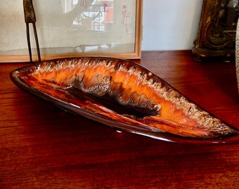 May include: A long, oval-shaped ceramic serving dish with a brown, orange, and white glaze. The dish is sitting on a wooden table.