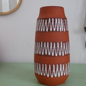 May include: A terracotta vase with white and brown wavy lines around the middle. The vase is sitting on a green surface.