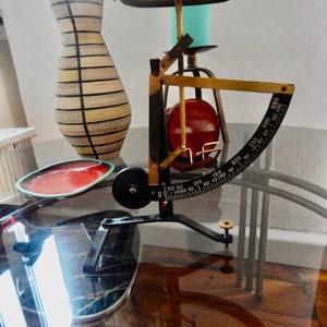 Könnte beinhalten: Eine antike Küchenwaage aus Messing und Schwarz mit einem roten und goldenen Zifferblatt. Die Waage steht auf einem Glastisch mit einem roten Teller und einer Vase mit einem gestreiften Muster.