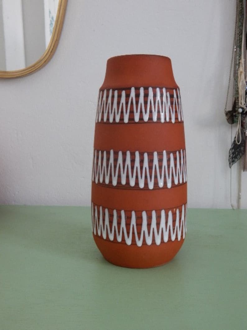 May include: A terracotta vase with white and brown wavy lines around the middle. The vase is sitting on a green surface.