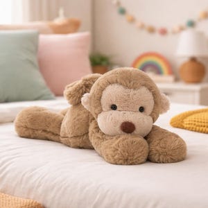 Puede incluir: Un juguete de peluche de mono marrón claro con una cara color crema y una nariz marrón. El animal de peluche está acostado sobre una colcha blanca. El mono tiene ojos pequeños y oscuros y una textura suave y peluda. El juguete está diseñado para niños.