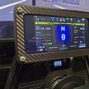 May include: A rectangular digital dashboard display mounted on a carbon fiber patterned frame with four silver screws. The screen shows racing telemetry data including a gear indicator set to N, speed, lap times, and weather information in a dark interface.