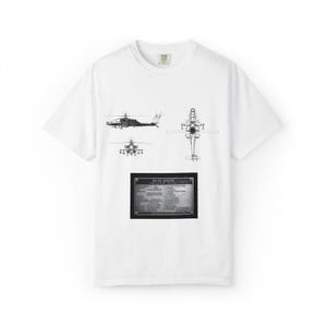 May include: White t-shirt with black line drawings of an AH-64 Apache helicopter from different perspectives. A rectangular plaque with text is below the drawings. The shirt has a crew neck and short sleeves.