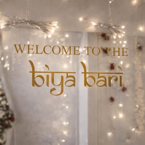 Pode incluir: Uma placa decorativa com as palavras "WELCOME TO THE biya bari" em letras douradas. A placa está colocada sobre um fundo de tecido branco macio, luzes de fadas e arranjos florais, criando uma atmosfera acolhedora e convidativa.