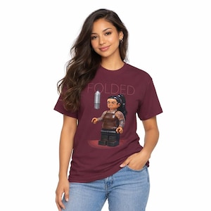 Puede incluir: Camiseta burdeos con la palabra "FOLDED" impresa en la parte superior. La camiseta presenta un gráfico de una figura de Lego sosteniendo un objeto plateado. La figura de Lego tiene tatuajes y lleva un top marrón y pantalones negros.