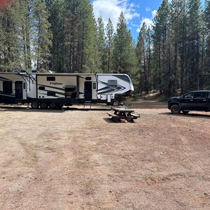 May include: A large white and black recreational vehicle with the word "Fuzion" on the side is parked on a dirt lot. A black pickup truck is parked nearby. A wooden picnic table sits in front of the RV.