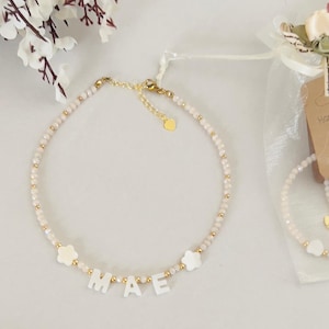 May include: A delicate necklace with the name "MAE" spelled out in white letters. The necklace features small, light-colored beads and gold accents, including a heart-shaped charm. Two white flower-shaped charms are also present.