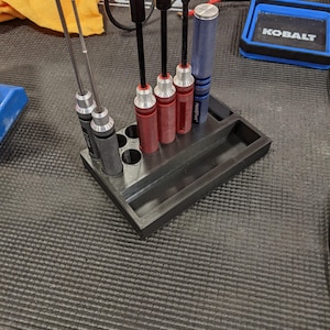 May include: A black 3D-printed tool organizer holding various screwdrivers and hex drivers. The tools have red, blue, and black handles. The organizer sits on a textured surface, with a blue Kobalt container visible in the background.