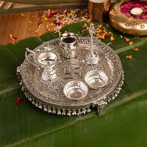 Könnte beinhalten: Ein silberfarbenes Puja-Thali-Set aus Metall mit einem verzierten runden Tablett, das ein zentrales Swastika-Symbol aufweist, sowie einer kleinen Glocke, einem Behälter mit Deckel, einer Öllampe mit Griff und zwei kleinen Schalen mit floralen Mustern.