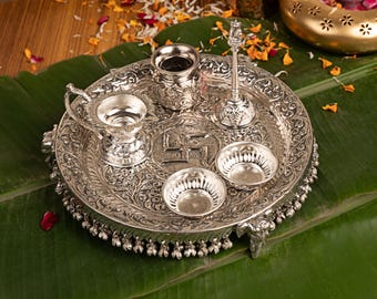 Può includere: Un set puja thali in metallo color argento composto da un vassoio circolare decorato con un simbolo svastica centrale, una piccola campana, un contenitore con coperchio, una lampada a olio con manico e due ciotole con motivi floreali e ciondoli.