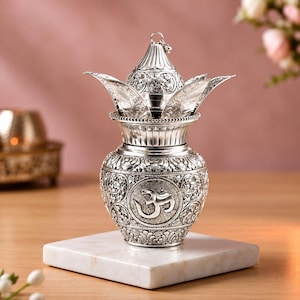 Könnte beinhalten: Ein silberfarbener Metall-Kalash-Topf mit einem geprägten Om-Symbol auf der Vorderseite. Das Gefäß hat einen bauchigen Körper mit detaillierten floralen Gravuren und einen spitzen Deckel mit kleiner Öse, umgeben von fünf dekorativen Blütenblättern.