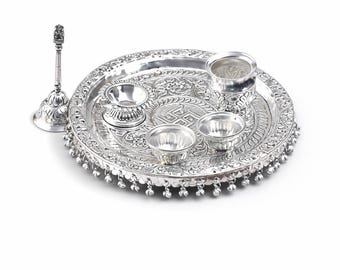 Peut inclure: Un ensemble de thali de puja en argent avec des motifs floraux et géométriques complexes. L'ensemble comprend un plateau rond, de petits bols, une tasse et un support décoratif. Le plateau a un bord perlé et un motif central. Les articles sont en métal argenté brillant.