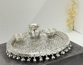 Peut inclure: Un plateau circulaire en métal argenté orné de motifs floraux en relief complexes et d'un symbole swastika central. Le plateau est agrémenté d'une frange de petites clochettes suspendues sur le bord et contient un petit pot et deux bols assortis.
