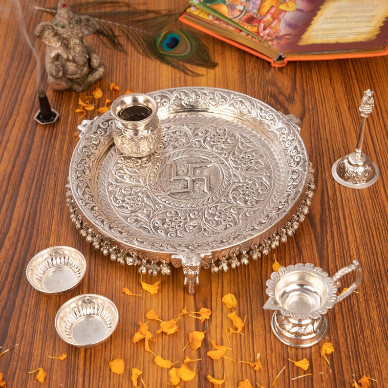 Peut inclure: Un ensemble de puja thali en m&eacute;tal argent&eacute; comprenant un grand plateau circulaire avec des gravures florales complexes et un symbole swastika central, accompagn&eacute; d'un petit pot, deux bols, une lampe &agrave; huile &agrave; poign&eacute;e et une cloche assortis.