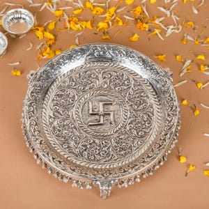 Peut inclure: Une assiette ronde en m&eacute;tal argent&eacute; orn&eacute;e d'un motif floral en relief complexe et d'un symbole swastika central. L'assiette pr&eacute;sente une bordure d&eacute;corative en relief et une s&eacute;rie de petites breloques m&eacute;talliques suspendues sur le bord ext&eacute;rieur.