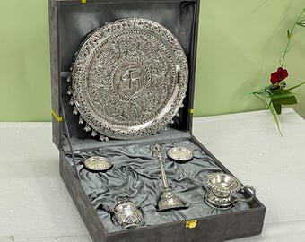 Peut inclure: Un ensemble de thali de puja argenté, comprenant un grand plateau orné avec un motif central, de petits bols, une cuillère et une tasse avec une poignée. L'ensemble est présenté dans une boîte doublée de velours gris avec un fermoir doré.