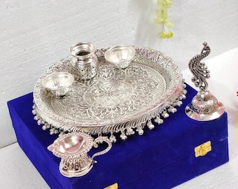 Peut inclure: Un ensemble de puja thali argenté, comprenant un plateau ovale orné de motifs détaillés, de petits bols et une figurine de paon. Les articles sont présentés sur une boîte en velours bleu, probablement à des fins cérémonielles. Le plateau mesure environ 25 cm de long.