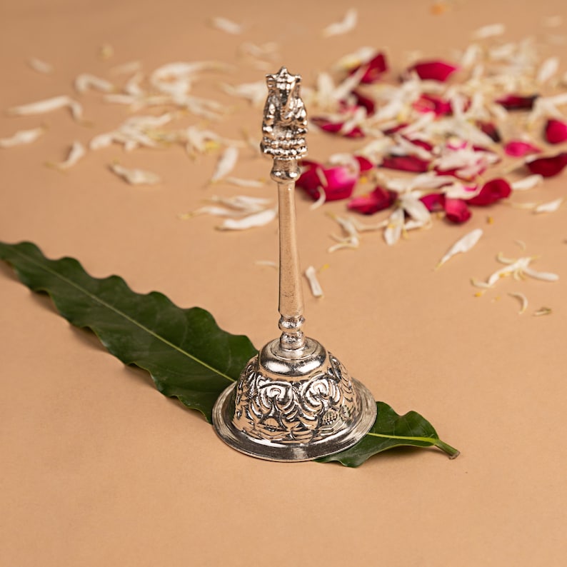 Peut inclure: Une cloche &agrave; main en m&eacute;tal argent&eacute; pr&eacute;sentant un motif floral orn&eacute; en relief sur la base et une petite figurine d&eacute;taill&eacute;e de la divinit&eacute; Ganesha au sommet du manche. La cloche poss&egrave;de une tige longue et fine ainsi qu'une base circulaire &eacute;vas&eacute;e.